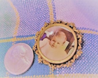 以下が含まれることがあります： 赤ちゃんが写った丸い肖像画が入った、ヴィンテージの金色のロケット。ロケットには装飾的な花の縁取りがあり、コインの隣にあります。背景は青と黄色のチェック柄の生地です。