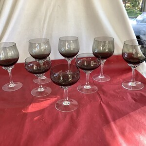 May include: Set of eight clear glass wine glasses with a dark gray tint. The glasses are arranged on a red tablecloth.