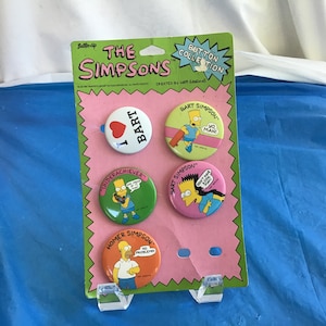 May include: A pink and white card with a pink background and white scalloped edges. The card features five round buttons with images of The Simpsons characters. The buttons feature Bart Simpson, Homer Simpson, and a heart with the word "Bart" on it. The text on the card reads "The Simpsons Button Collection" and "Created by Hatt Galleries".
