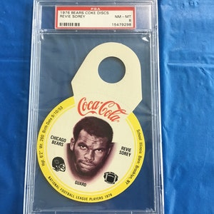 May include: A yellow and brown Coca-Cola disc featuring a photo of football player Revie Sorey, a guard for the Chicago Bears. The disc is a 1976 National Football League Players collectible. The text "Coca-Cola" is printed in red on the disc.