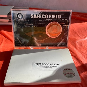 May include: A clear plastic case containing a coin and a card. The coin features an image of a stadium and the text "HOME OF THE SEATTLE MARINERS". The card has the text "SAFECO FIELD" and "MLB MARINERS STADIUM COIN/CARD".