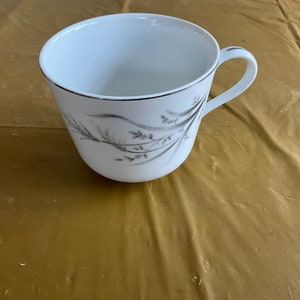 May include: White teacup with a handle and a delicate floral design in gray. The rim of the cup is accented with a thin silver band. The cup is sitting on a gold-colored surface.