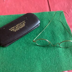 May include: A brown leather glasses case with the text "CLIFFORD E. SMITH, B.S., M.D. ROBB V. SMITH, B.A., M.D. OCULISTS and AURISTS DE KALB, ILLINOIS" printed on the front. A pair of gold-rimmed round glasses with thin wire frames are lying on a green surface.