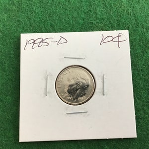 May include: A 1995-D dime coin in a white cardboard holder. The coin features a profile of a person and the word "LIBERTY". The holder has handwritten text that reads "1995-D" and "10¢". The background is green.