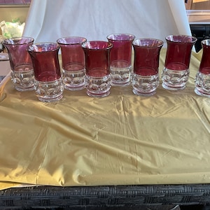 May include: A set of nine vintage drinking glasses. The glasses have a clear, patterned base and a deep red upper portion. The glasses are arranged on a gold-colored surface.