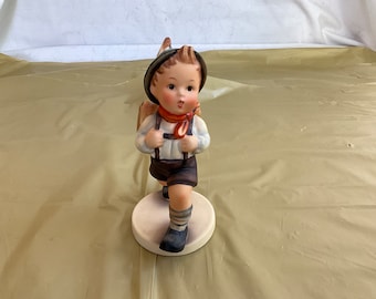 Puede incluir: Una figurita de porcelana de un niño que lleva un sombrero verde, una bufanda roja y una camisa azul y blanca. Lleva una mochila marrón y tiene una cinta roja y blanca atada alrededor del cuello. La figurita está de pie sobre una base blanca.