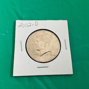 May include: A 2002-D Kennedy half dollar coin, displayed in a protective holder. The coin features a profile of John F. Kennedy and the inscriptions "IN GOD WE TRUST" and "LIBERTY". The coin is a golden color.