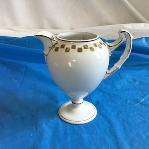 May include: White ceramic creamer with a gold trim and a geometric pattern around the top. The creamer has a handle and a pedestal base.