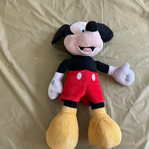 May include: A plush Mickey Mouse toy with black fur, red shorts, and yellow shoes. The toy has a big smile and is lying on a tan surface.