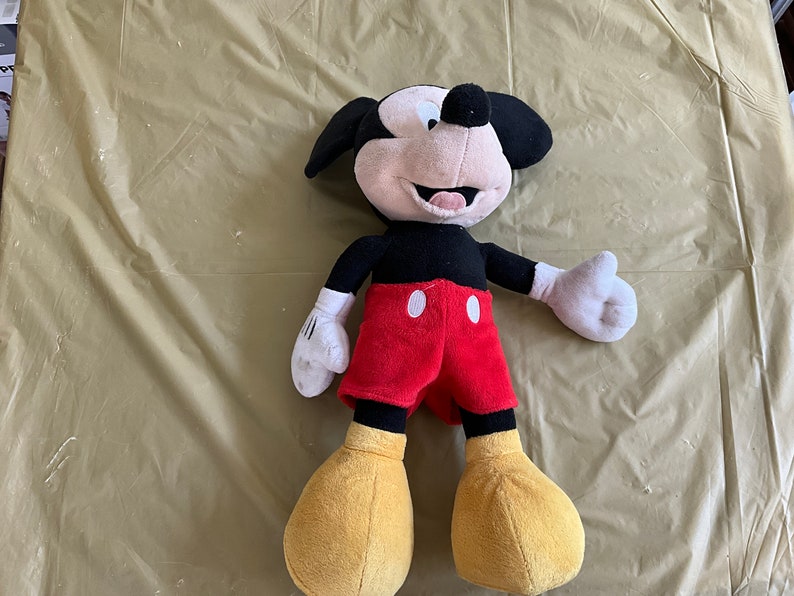 May include: A plush Mickey Mouse toy with black fur, red shorts, and yellow shoes. The toy has a big smile and is lying on a tan surface.