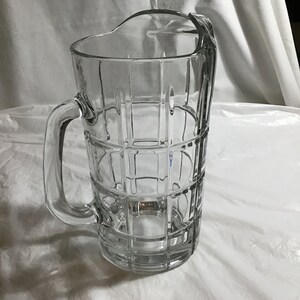 May include: A clear glass pitcher with a geometric pattern. The pitcher has a handle and a wide mouth.