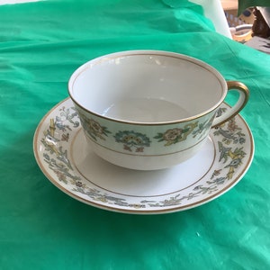 May include: A white teacup and saucer with a floral pattern in shades of green, yellow, and blue. The cup has a gold rim and the saucer has a gold rim and a gold band around the edge.
