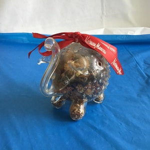 May include: A clear glass elephant ornament with a red ribbon that says "Neiman Marcus". The elephant is filled with dried flowers and other botanicals.