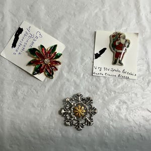 May include: Three decorative brooches. One is a poinsettia with red, green, and gold enamel and rhinestones. Another is a vintage Santa Claus brooch. The third is a silver-tone snowflake with a gold center.