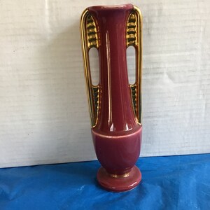 May include: A small, burgundy ceramic vase with gold and green accents. The vase has two handles and a rounded base.