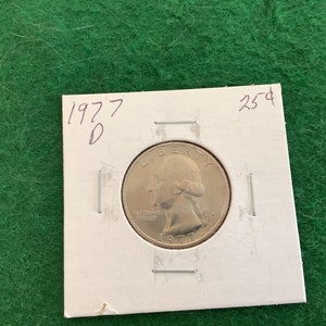 May include: A silver 1977-D quarter dollar coin with the inscription "IN GOD WE TRUST" and "LIBERTY" on the obverse side. The coin is displayed on a white card with the year "1977" and the letter "D" written in black ink. The card also has the inscription "25¢" written in black ink.