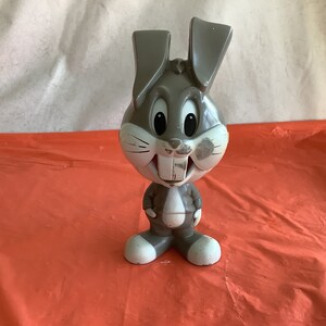 May include: A gray plastic Bugs Bunny figurine with a white face and large ears. The figurine is standing on a red surface.