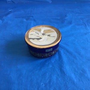 May include: A round blue and gold trinket box with a 24KT gold lid featuring a white and gold image of two cranes in flight.