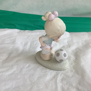 May include: A porcelain figurine of a girl in a blue and white soccer uniform, with a pink bow in her hair, looking down at a soccer ball.