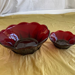 May include: Two red glass bowls with a scalloped edge. The larger bowl is in the background and the smaller bowl is in the foreground.