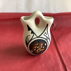 May include: A white ceramic vase with a black and orange geometric design. The vase has a rounded shape and a small opening at the top.