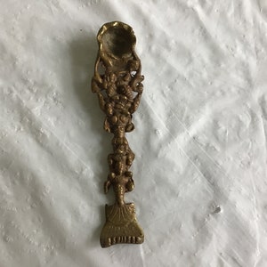 May include: A gold-colored metal spoon with intricate floral designs and a rounded bowl. The spoon has a long, slender handle with a decorative design.