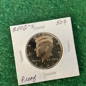 May include: A silver US half dollar coin with the profile of John F. Kennedy on the obverse. The coin is dated 2002 and has the inscription "In God We Trust". The coin is displayed on a white card with the text "2002-S" and "Proof" written on it.