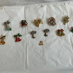 May include: A collection of 12 gold, silver, and red Christmas bell pins with green holly accents. Some of the bells have rhinestones and glitter.