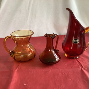 May include: Three glass pitchers in amber and red. The amber pitchers have a rounded body and a flared lip. The red pitcher has a tall, slender body and a flared lip. All three pitchers have a handle.