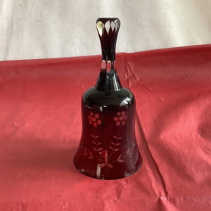 May include: A dark red glass bell with a floral design. The bell has a clear glass handle and is etched with a floral pattern.
