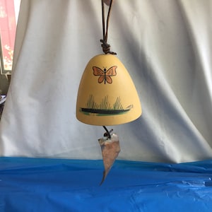 May include: A yellow ceramic bell with a painted orange butterfly and green grass design. The bell is hanging from a brown leather cord.