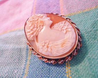 Puede incluir: Un broche de camafeo con un retrato tallado de una mujer con un diseño floral. El camafeo está engastado en un marco dorado con un borde de perlas.