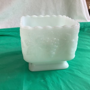 May include: White milk glass bowl with a square shape and a scalloped rim. The bowl has a raised floral design and a pedestal base.