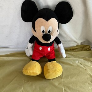 May include: A plush Mickey Mouse toy with black fur, large ears, and a red and white outfit. The toy is sitting on a yellow surface.