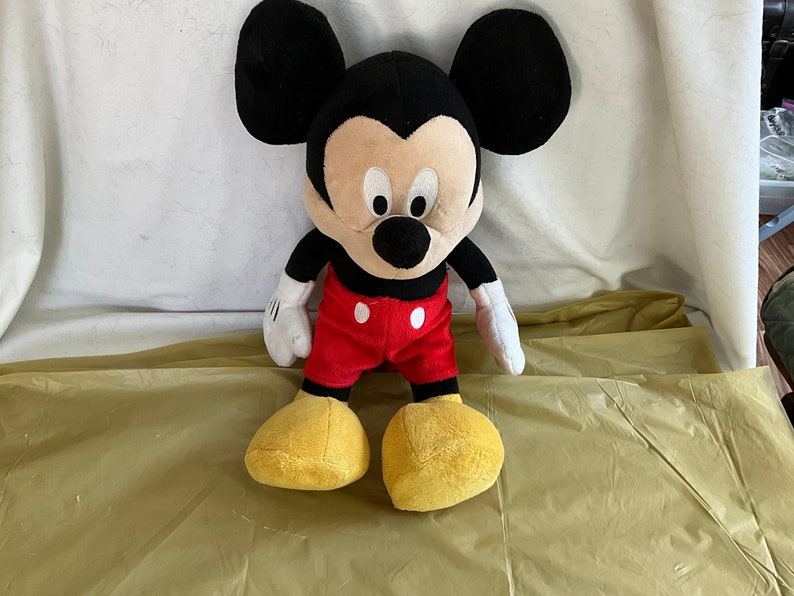 May include: A plush Mickey Mouse toy with black fur, large ears, and a red and white outfit. The toy is sitting on a yellow surface.