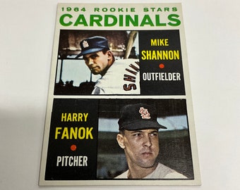 May include: 1964 Rookie Stars Cardinals baseball card featuring Mike Shannon, Outfielder and Harry Fanok, Pitcher. The card has a red dot next to each player's name.