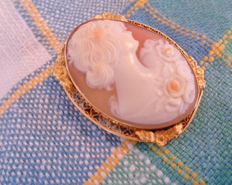 May include: A cameo brooch featuring a carved portrait of a woman with flowers, set in a gold filigree frame.