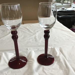 May include: Two clear glass wine glasses with burgundy red stems and bases. The glasses are empty and sitting on a white tablecloth.