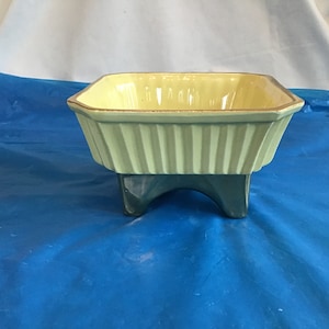 May include: A square ceramic planter with a light green body and a yellow rim. The planter has four short legs and is designed to hold a small plant.