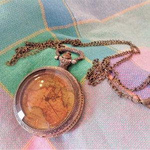 May include: A gold-toned pocket watch with a map design on the face, attached to a gold chain. The watch is lying on a pink and green checkered fabric.