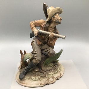 May include: A ceramic figurine depicting a hunter kneeling with a rifle, aiming at a rabbit. The hunter wears a brown jacket, hat, and boots. The base features sculpted foliage and a rabbit. The figurine is approximately 10 inches tall.