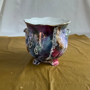 May include: A ceramic planter with a scalloped rim and two handles. The planter is decorated with a colorful abstract design in shades of purple, blue, green, and pink. The planter has three feet and is made of white porcelain.
