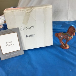 May include: A small, red, ceramic figurine of a Persian unicorn with gold and blue accents. The unicorn is standing on a red base. The figurine is packaged in a white box with the text "Persian Unicorn" and "The Franklin Mint" on a gray card.