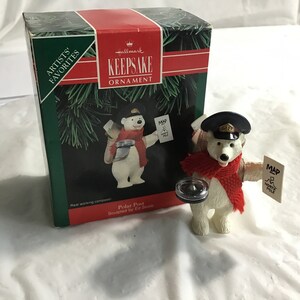 May include: A Hallmark Keepsake ornament featuring a polar bear dressed as a postal worker. The bear wears a black hat and red scarf, holding a map that reads "North Pole." The ornament is next to its original box.