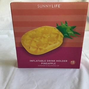 May include: A pink and orange box with a yellow inflatable pineapple drink holder. The box says "Inflatable Drink Holder Pineapple Summer is blowing up" and has a +6 years age recommendation.