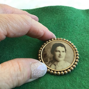 May include: A gold-toned locket with a beaded border, containing a black and white photograph of a woman with dark hair. The woman is wearing a white blouse with a lace collar.
