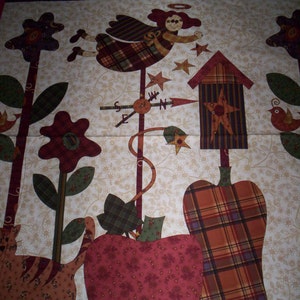 May include: A patchwork quilt block with a whimsical autumn theme. It features a red and green plaid angel, a birdhouse, a pumpkin, an apple, a cat, and a flower. The block is set against a cream background with a floral pattern.