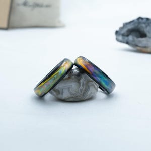 Peut inclure: Deux bagues irisées avec un effet multicolore de type nacre, posées sur une pierre grise à motifs. Les bagues présentent un spectre de couleurs, dont l'or, le bleu et le vert. Le fond est blanc doux, avec une étiquette floue et une roche.