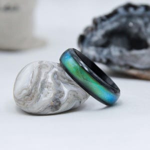 May include: A colour-changing mood ring with a black band, showing green and blue hues. The ring sits on a grey and white stone, with a blurred rock in the background. The ring's colour shifts according to the wearer's mood.