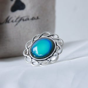 May include: A silver-toned ring with an oval, color-changing stone in shades of blue and green. The ring has a decorative, openwork design around the stone. A gray bag with the word "Miltraas" is in the background.